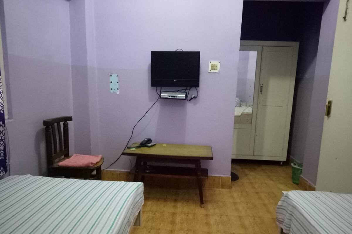 Una habitación con dos camas, un banco, un televisor montado en la pared y una puerta que da a otra estancia.