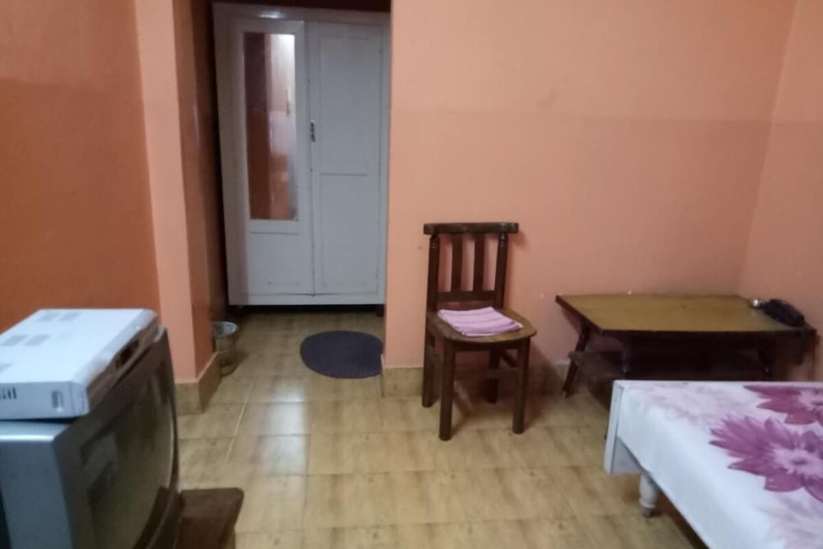 Una habitación con televisión, una silla de madera, una mesita pequeña y una cama con colcha estampada.