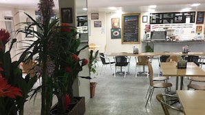 Ein Restaurantinneres mit einer langen Theke, mehreren Tischen und Stühlen. Entlang der Wand stehen üppige Pflanzgefäße mit roten und violetten Blumen.