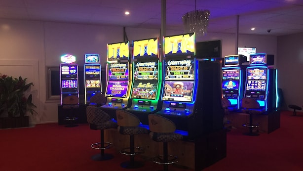 Eine Reihe von hell beleuchteten Spielautomaten in einem schwach beleuchteten Raum mit rotem Teppich und Barhocker.