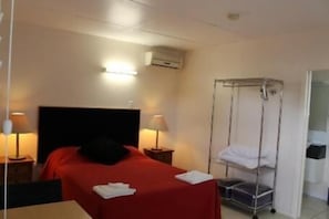 Habitación de hotel con cama, dos lámparas de noche, un equipo de aire acondicionado, un perchero con toallas y una puerta que da a otra habitación.