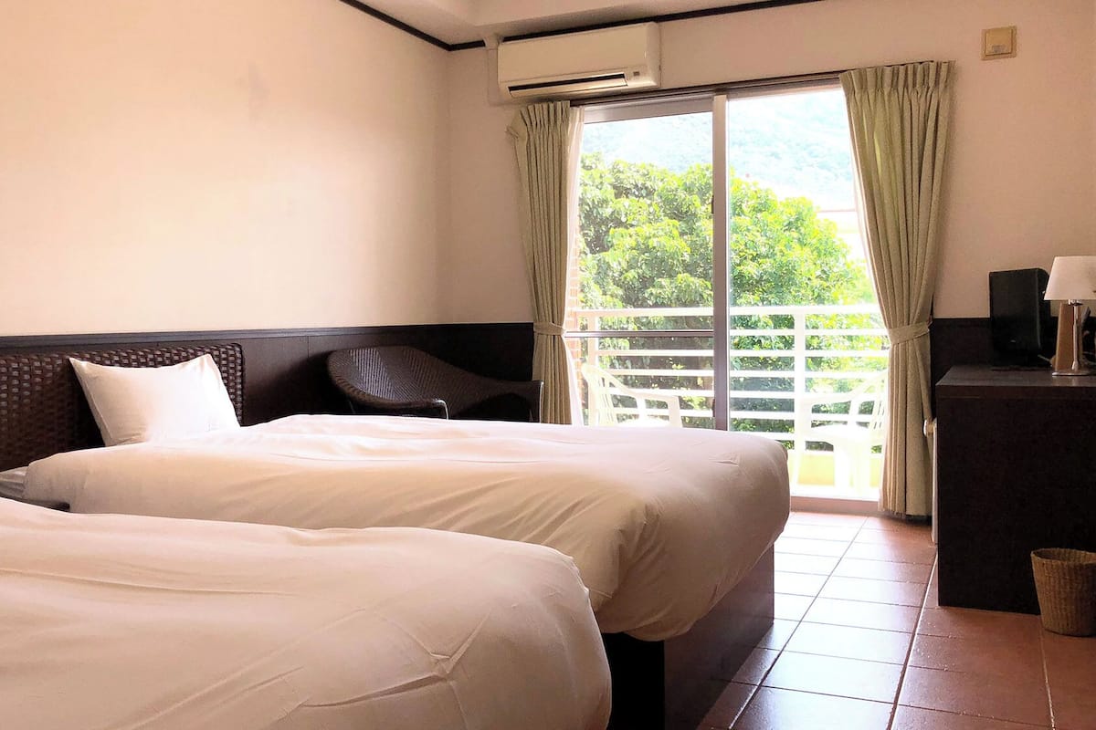 Une chambre d’hôtel avec deux lits, une table de chevet, une lampe, une télévision et un balcon donnant sur un paysage verdoyant.
