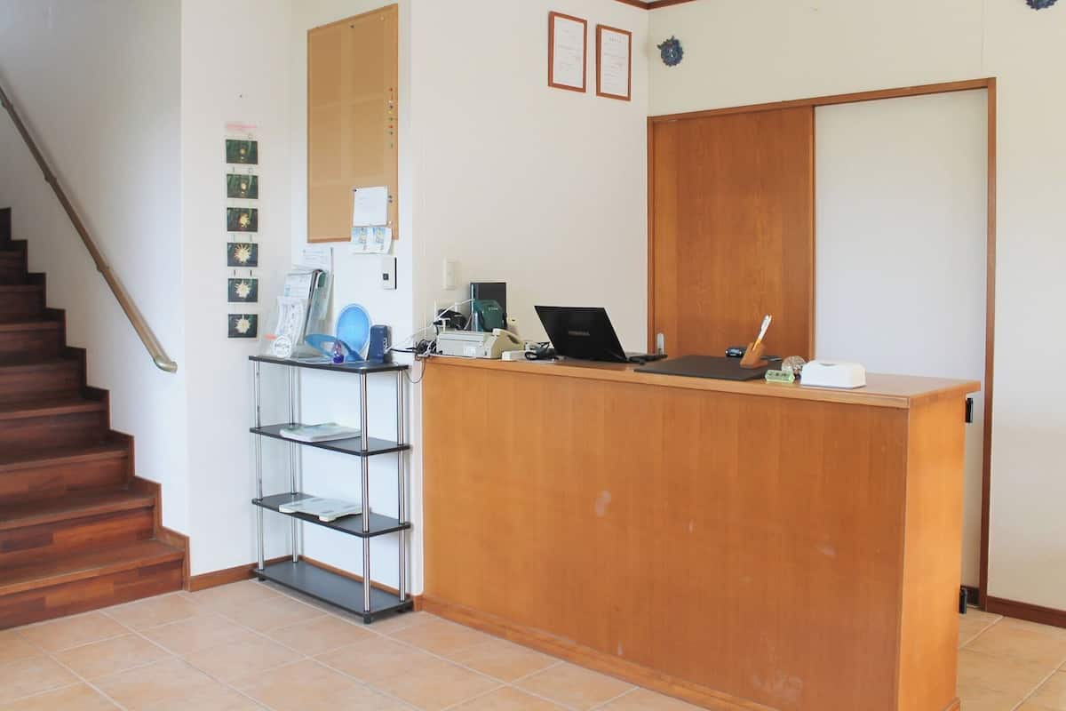 Un comptoir d’accueil avec un ordinateur portable, un téléphone et une petite étagère contenant divers objets. Un escalier se trouve à gauche et une porte à droite.