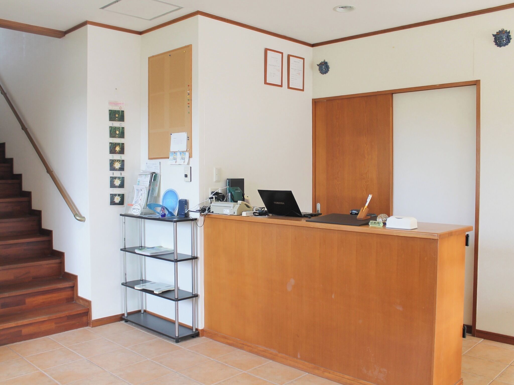 A reception desk with a laptop, a telephone, and a small shelf with items. There is a staircase to the left and a door to the right.