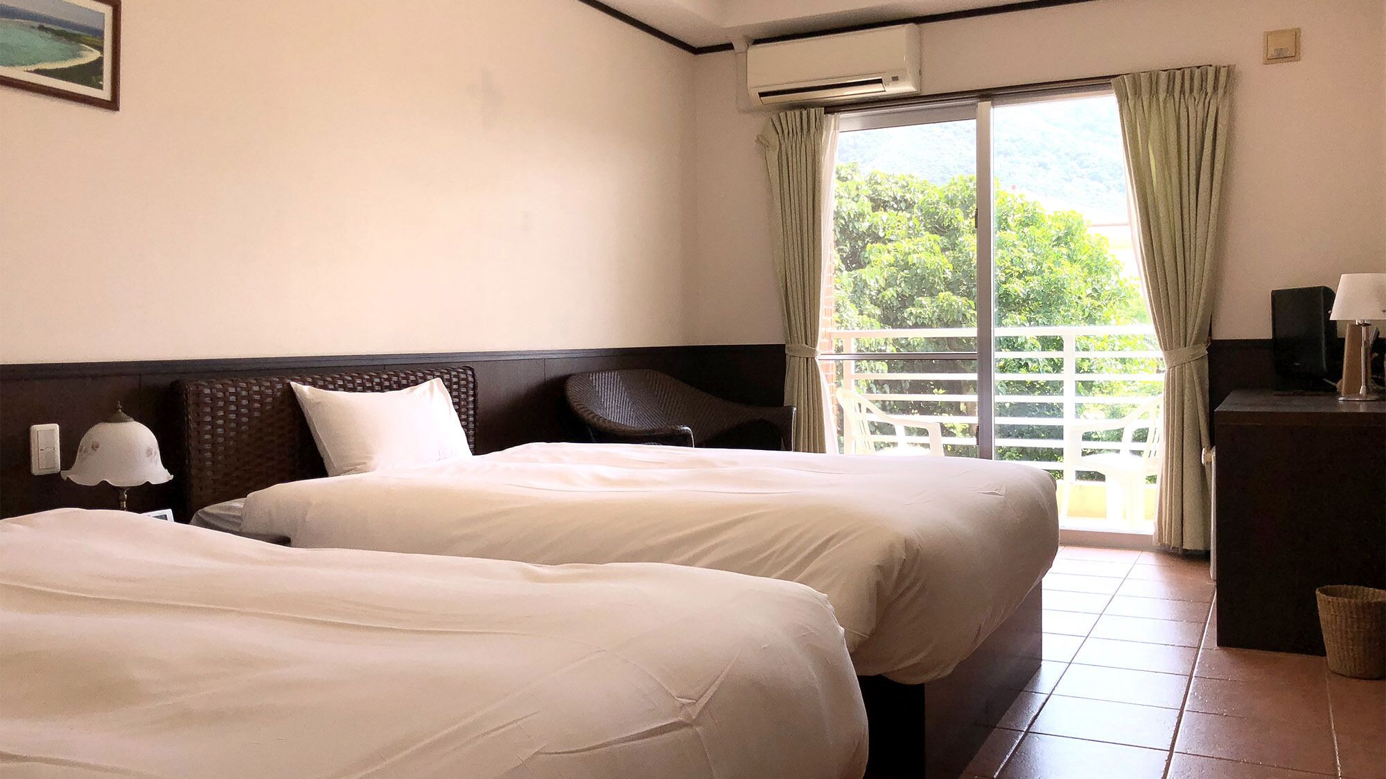 A hotel room with two beds, a nightstand with a lamp, a window with curtains, and a view of greenery outside.