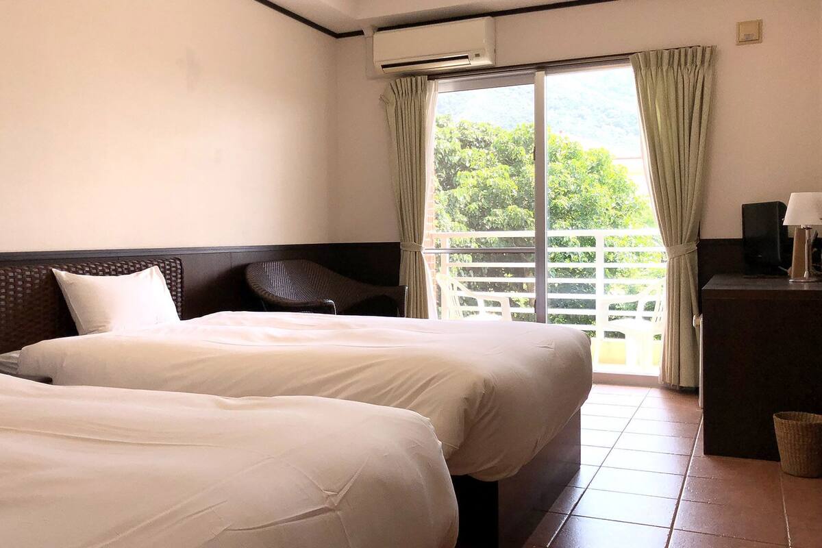 Une chambre d’hôtel avec deux lits, une table de chevet avec une lampe, une fenêtre avec des rideaux et une vue sur la verdure.