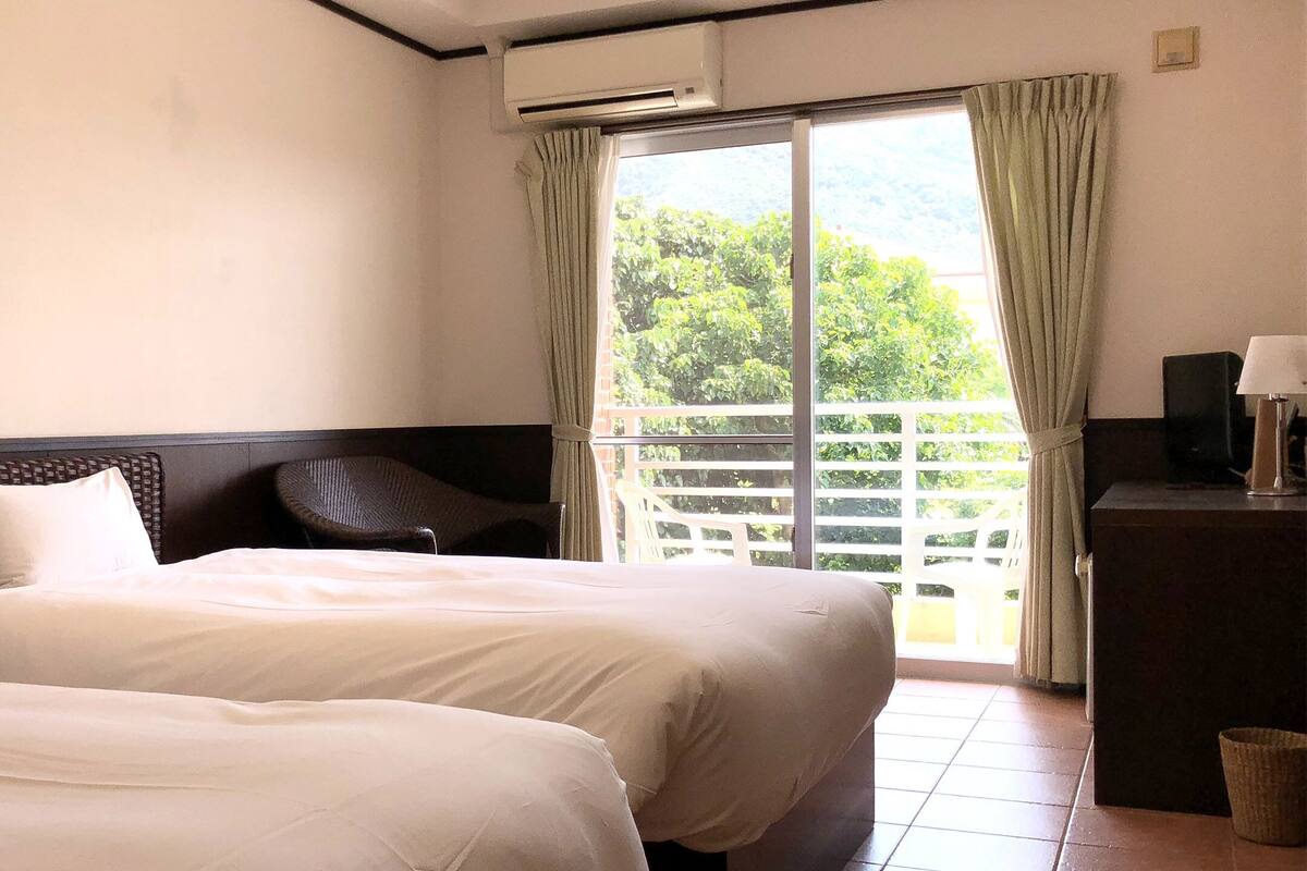 Une chambre d’hôtel avec deux lits, un balcon donnant sur un espace verdoyant et un bureau avec une lampe.