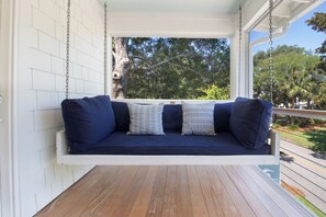 A porch swing with a dark blue cushion and patterned pillows, wooden flooring, and a view of trees and a clear sky. - Serene and Completely Renovated!~ Heron House (Folly Beach)