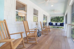 Une véranda couverte avec des chaises en bois et un fauteuil à bascule, des murs blancs et une vue sur la verdure.