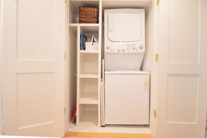 A laundry room with a washing machine and dryer, shelves with folded clothes, and a basket. - Serene and Completely Renovated!~ Heron House (Folly Beach)