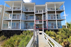 A multi-story beachfront building with balconies, a wooden deck, and surrounding vegetation.