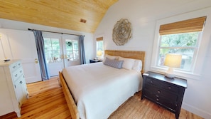 A bedroom with a wooden ceiling, a large bed with white bedding, a nightstand with a lamp, a dresser, and a window with a view of greenery.