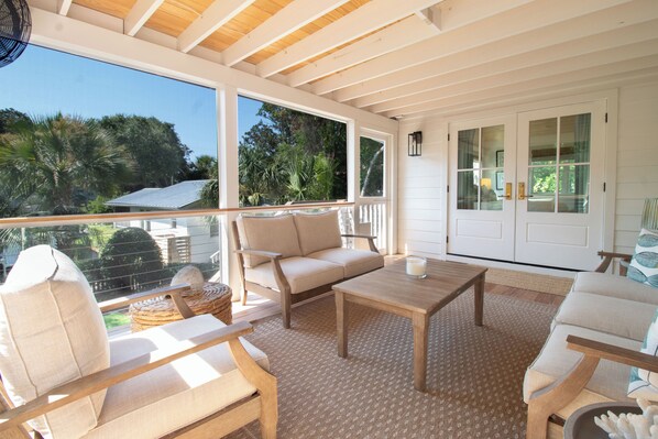A covered outdoor seating area with a sofa, chairs, and a table, overlooking a garden and a house.