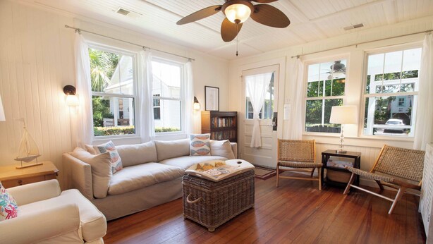 A spacious living room with a large sofa, a wicker ottoman, and two wicker chairs. There are two windows with curtains, a ceiling fan, and a bookshelf. - One house plus 2 cottages on the same lot~Family Houses (Folly Beach)