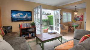 A cozy living room with a sofa, a coffee table, and a bookshelf. There is a painting on the wall and a window with blinds.