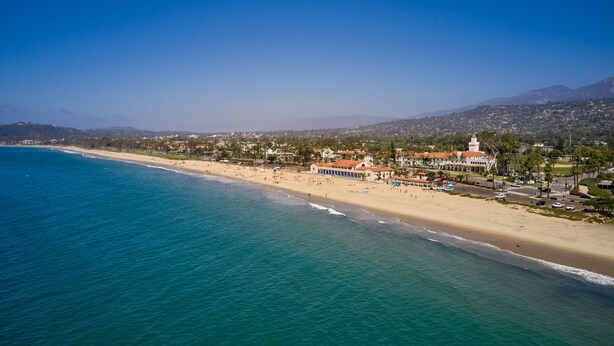 An aerial view of a coastal town with a beach, buildings, and a mountain range in the background.