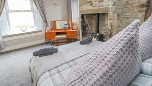 A bedroom with a stone fireplace, a wooden dresser, a bed with a plaid blanket, and a window with curtains. - 1 Braeside (Bishop Auckland)