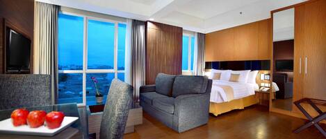A hotel room with a large bed, a sofa, a TV, and a dining area with apples on a table.