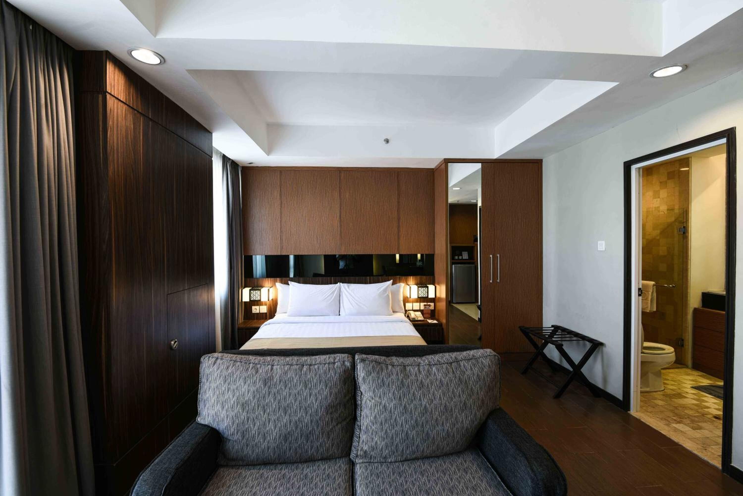 A hotel room with a bed, bedside tables, a sofa, a wardrobe, and a bathroom visible through an open door.