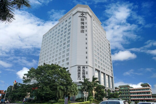 A tall, modern hotel with a prominent sign in a foreign script, surrounded by lush greenery and other buildings. - Rivan Hotel Longgang Shenzhen (Shenzhen)