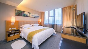 A hotel room with a large bed, a bedside table with a lamp, a desk with a chair, a large mirror, and a painting on the wall. - Rivan Hotel Longgang Shenzhen (Shenzhen)