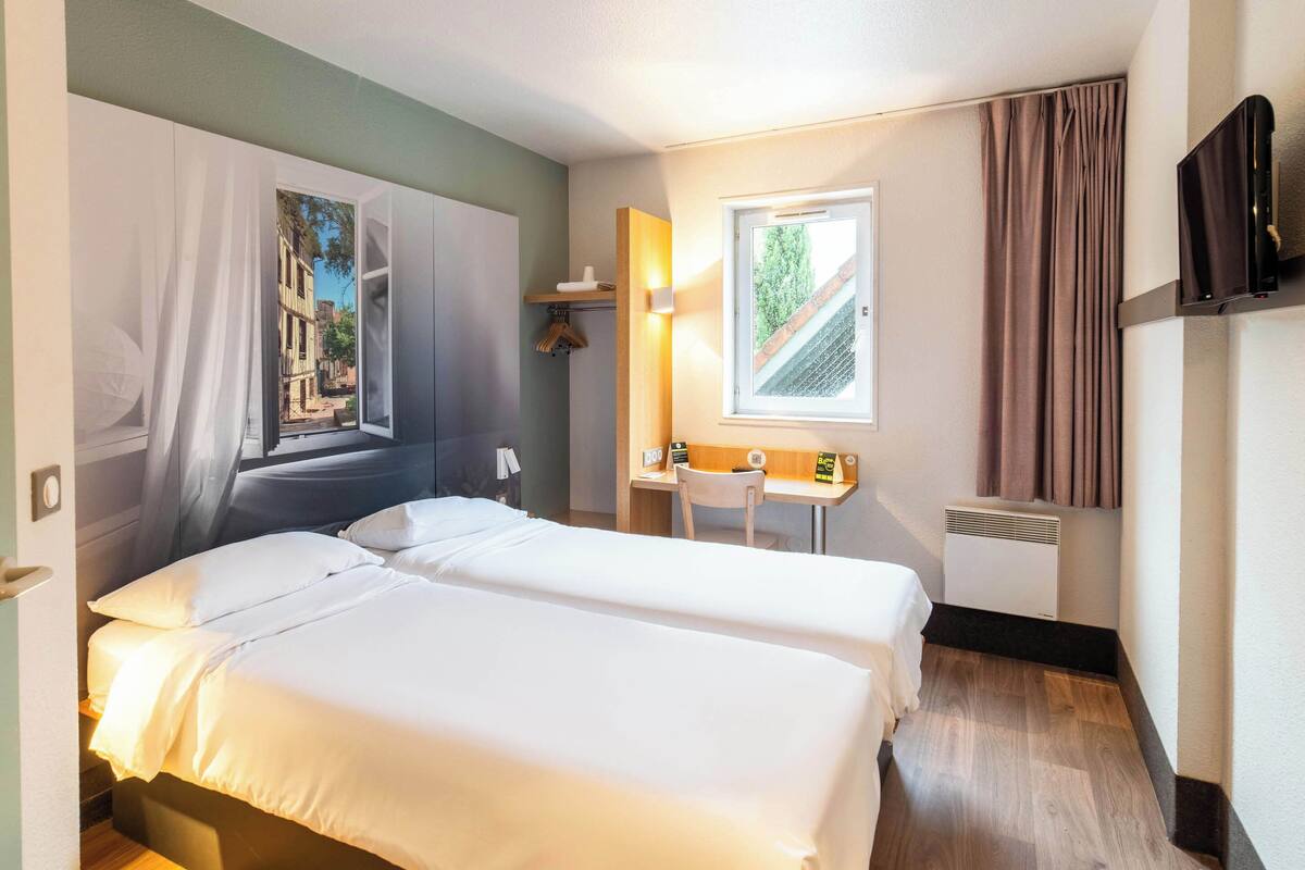 Une chambre d’hôtel avec deux lits, un bureau, une chaise, une télévision et une fenêtre avec des rideaux.