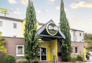 A B&B hotel with a yellow facade, two tall cypress trees, and a sign above the entrance. - B&B Hotel Limoges 1 (Limoges)