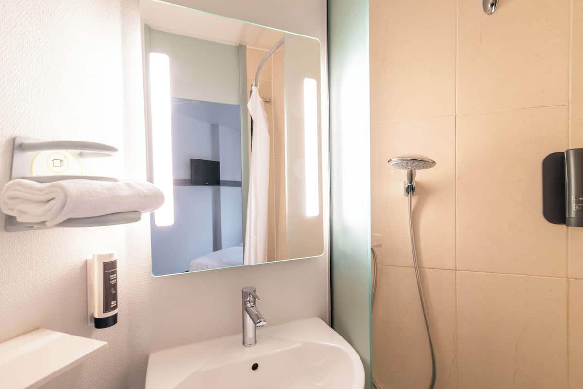 Une salle de bain avec un lavabo blanc, un miroir, un porte-serviettes, une douchette à main et un panneau de commande fixé au mur.
