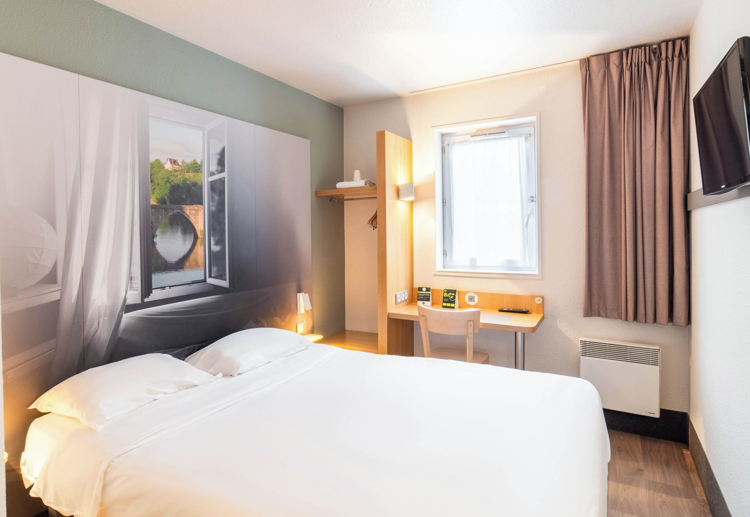 A hotel room with a bed, a desk, a chair, a window with curtains, a TV, and a radiator.
