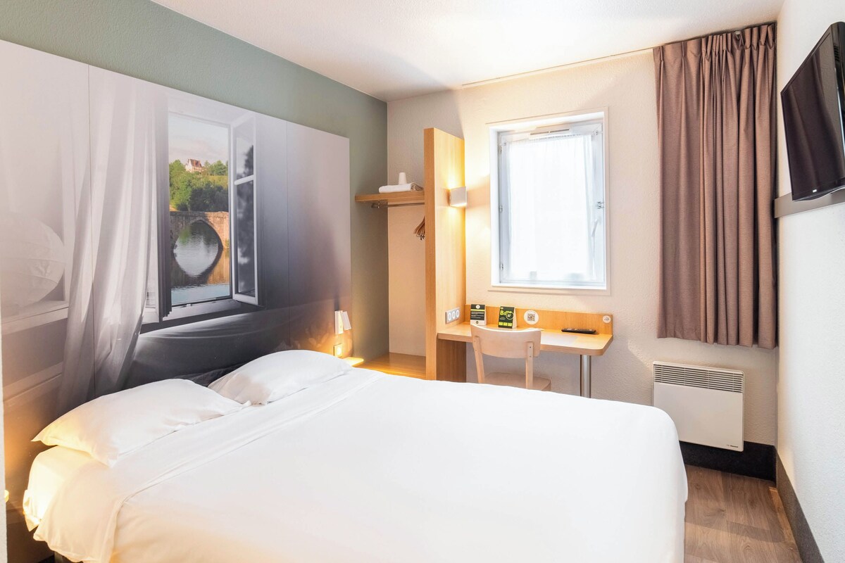 A hotel room with a bed, a desk, a chair, a window with curtains, a TV, and a radiator.