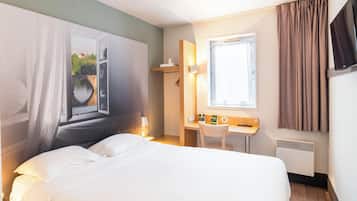 A hotel room with a bed, a desk, a chair, a window with curtains, a TV, and a radiator.