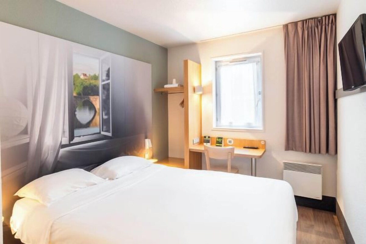 A hotel room with a bed, a desk, a chair, a window, and a TV.