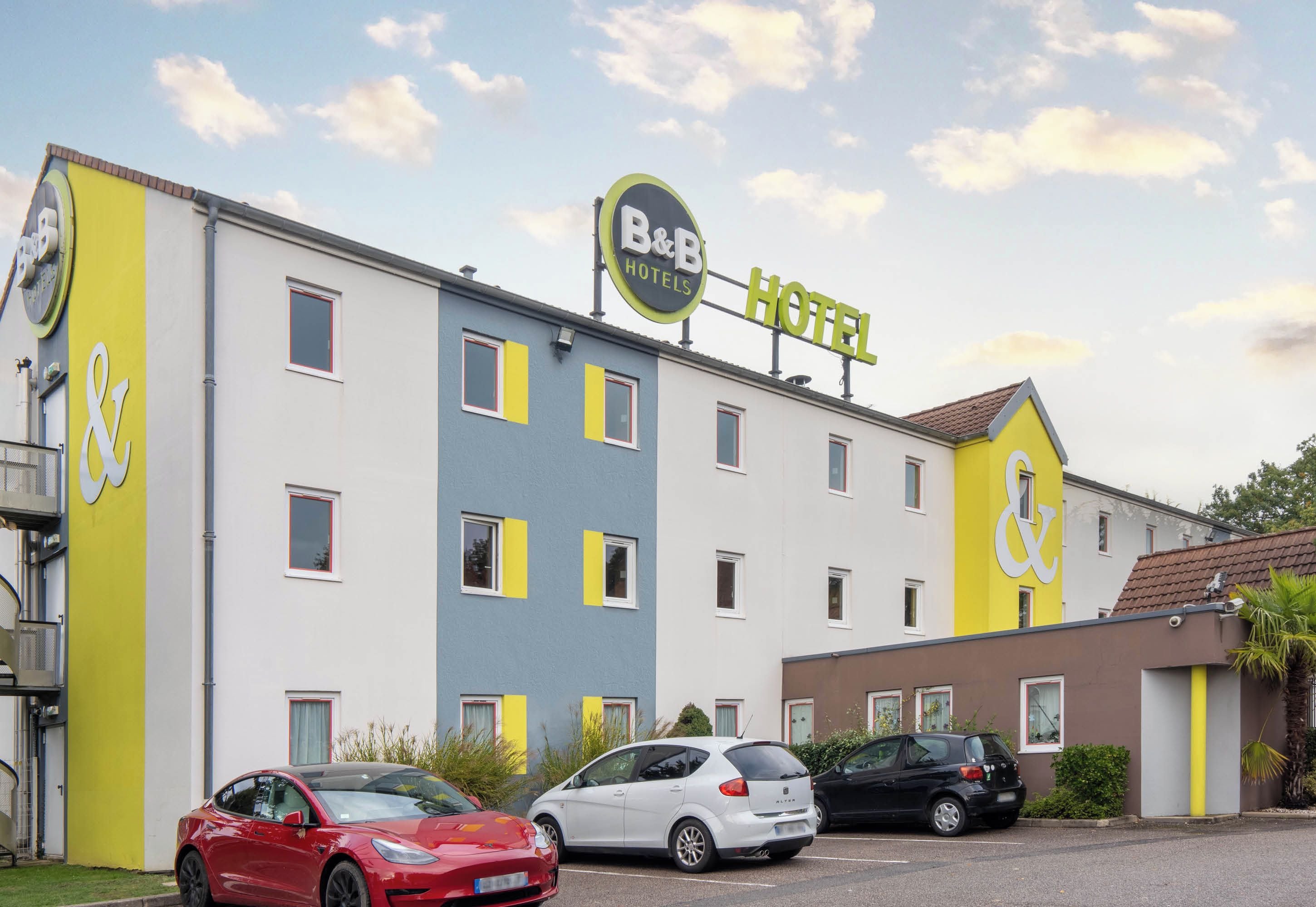 Un hotel B&B con diseño moderno, que destaca por un letrero llamativo, numerosas ventanas y un estacionamiento con varios autos.