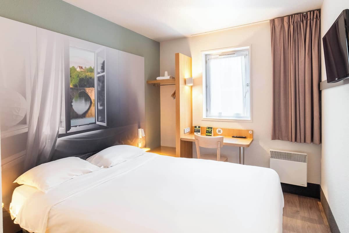 Une chambre d’hôtel équipée d’un lit, d’un bureau, d’une chaise, d’une fenêtre avec des rideaux, d’une télévision et d’un radiateur.