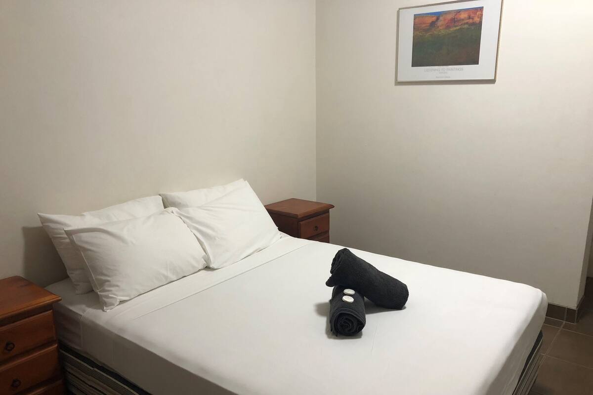 A neatly made bed with white linens, a black towel, and a pillow. There is a wooden nightstand with a drawer and a framed picture on the wall.