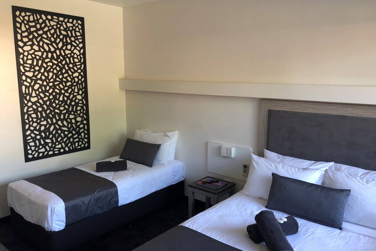 A hotel room with two beds, a decorative wall panel, a bedside table with a book, and a power outlet.