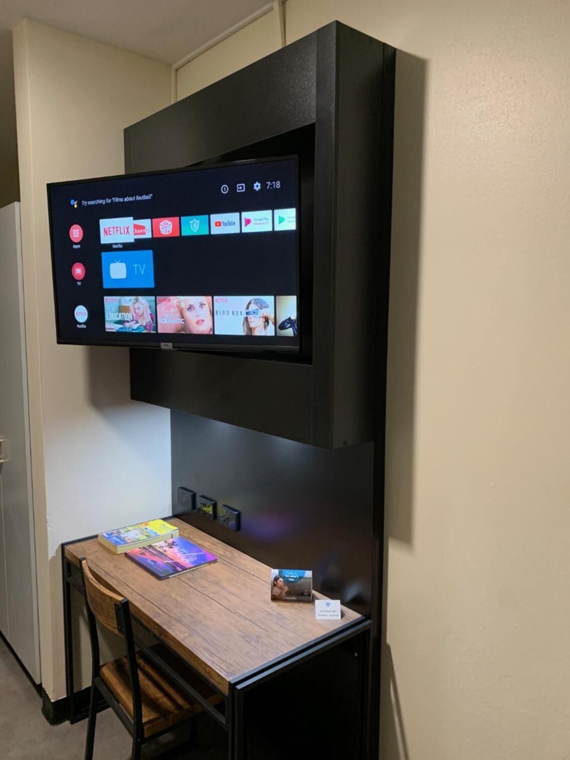 Een flatscreen televisie bevestigd aan een zwarte muur boven een houten tafel met boeken en een klein doosje.