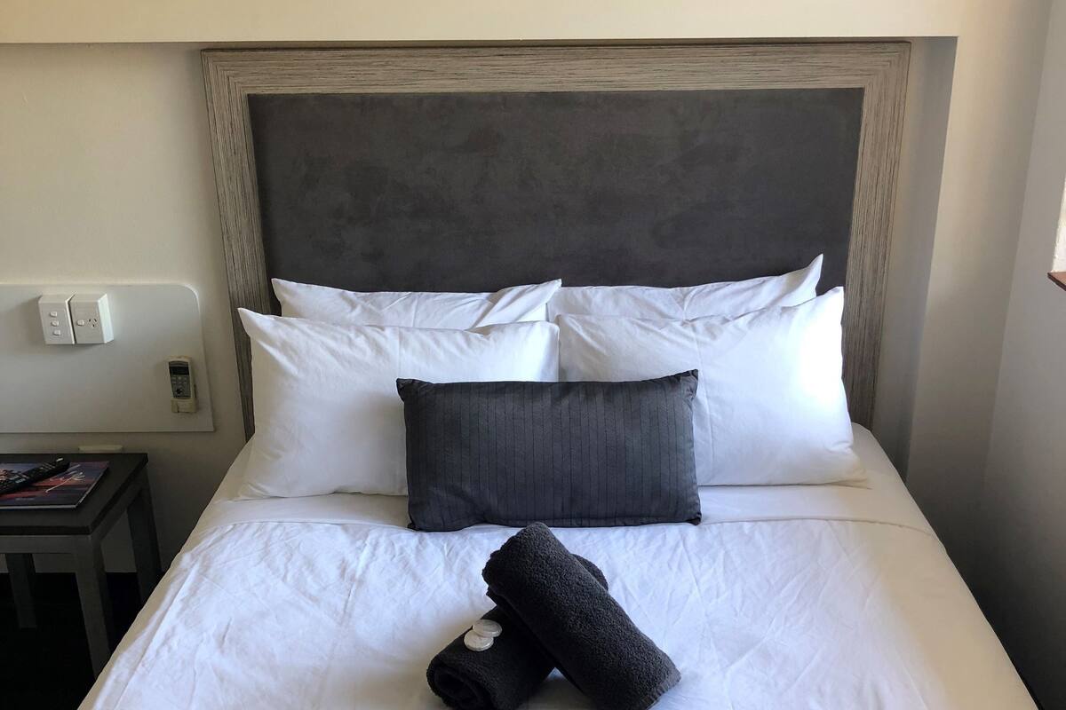 A neatly made bed with white and dark pillows, a folded towel, and a bedside table with a remote control.