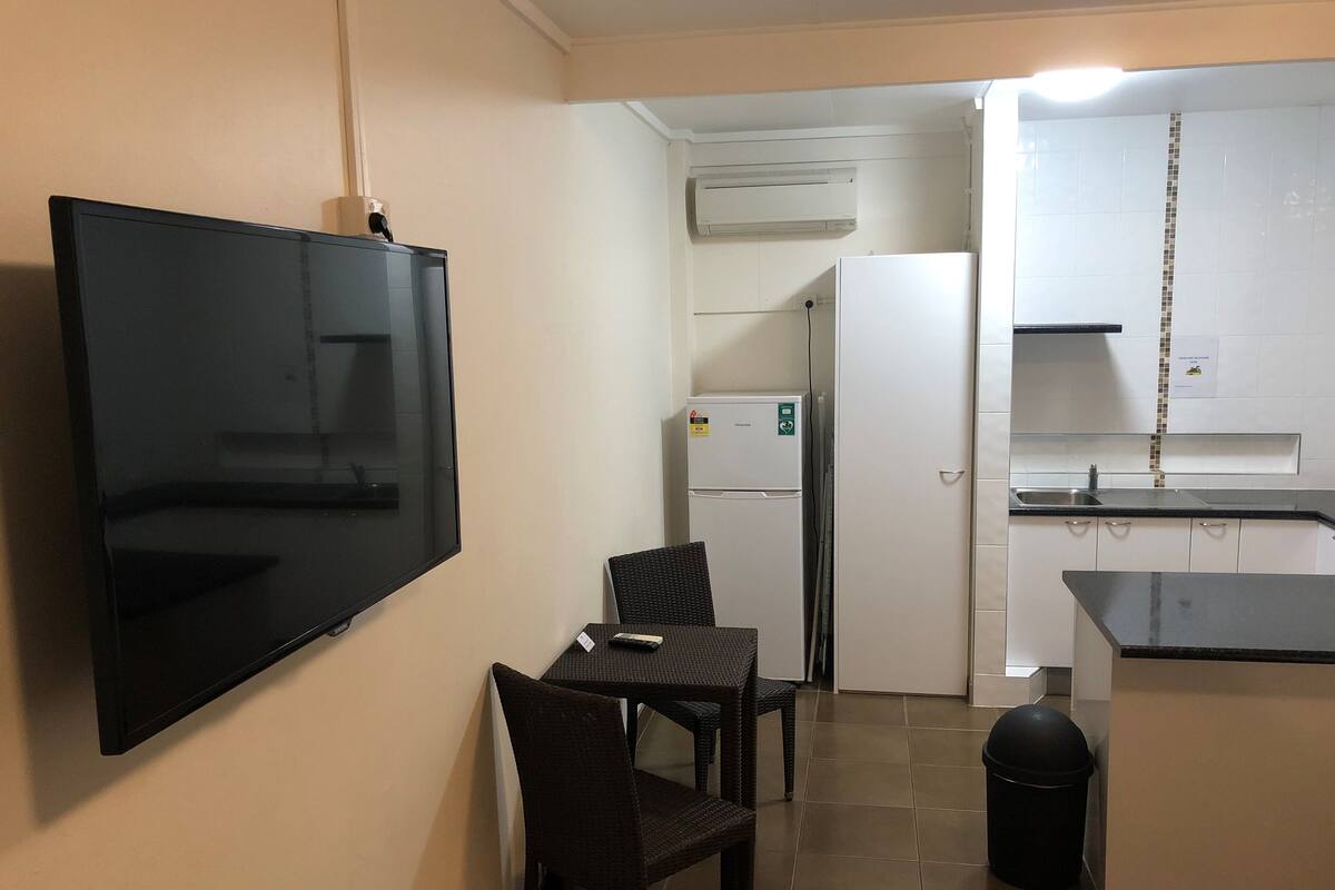 A compact kitchen with a flat-screen TV mounted on the wall, a refrigerator, and a small dining area with a table and chairs.