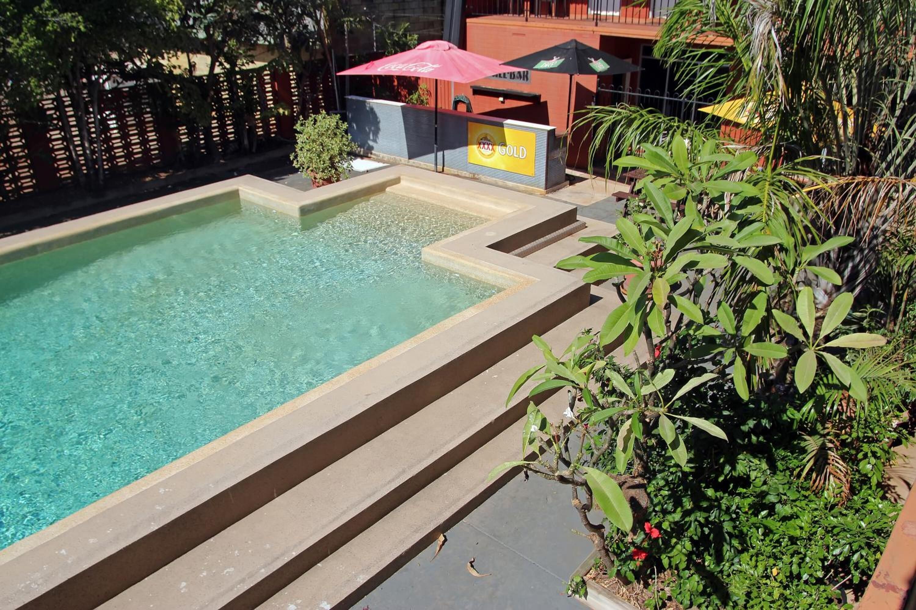 A swimming pool with steps leading into it, surrounded by greenery and a building with a 'Gold' sign.