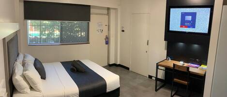A modern hotel room with a bed, a desk, a television, and a window with a view of trees.