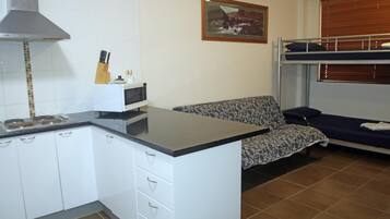 Una cucina compatta con mobili bianchi, un forno a microonde e un fornello. C'è un divano con motivo decorativo e un letto a castello con finestra.