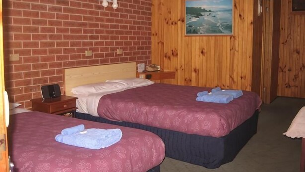 A room with two beds, wooden walls, a painting, and a small table with a lamp. - Orbost Country Road Motor Inn (Orbost)