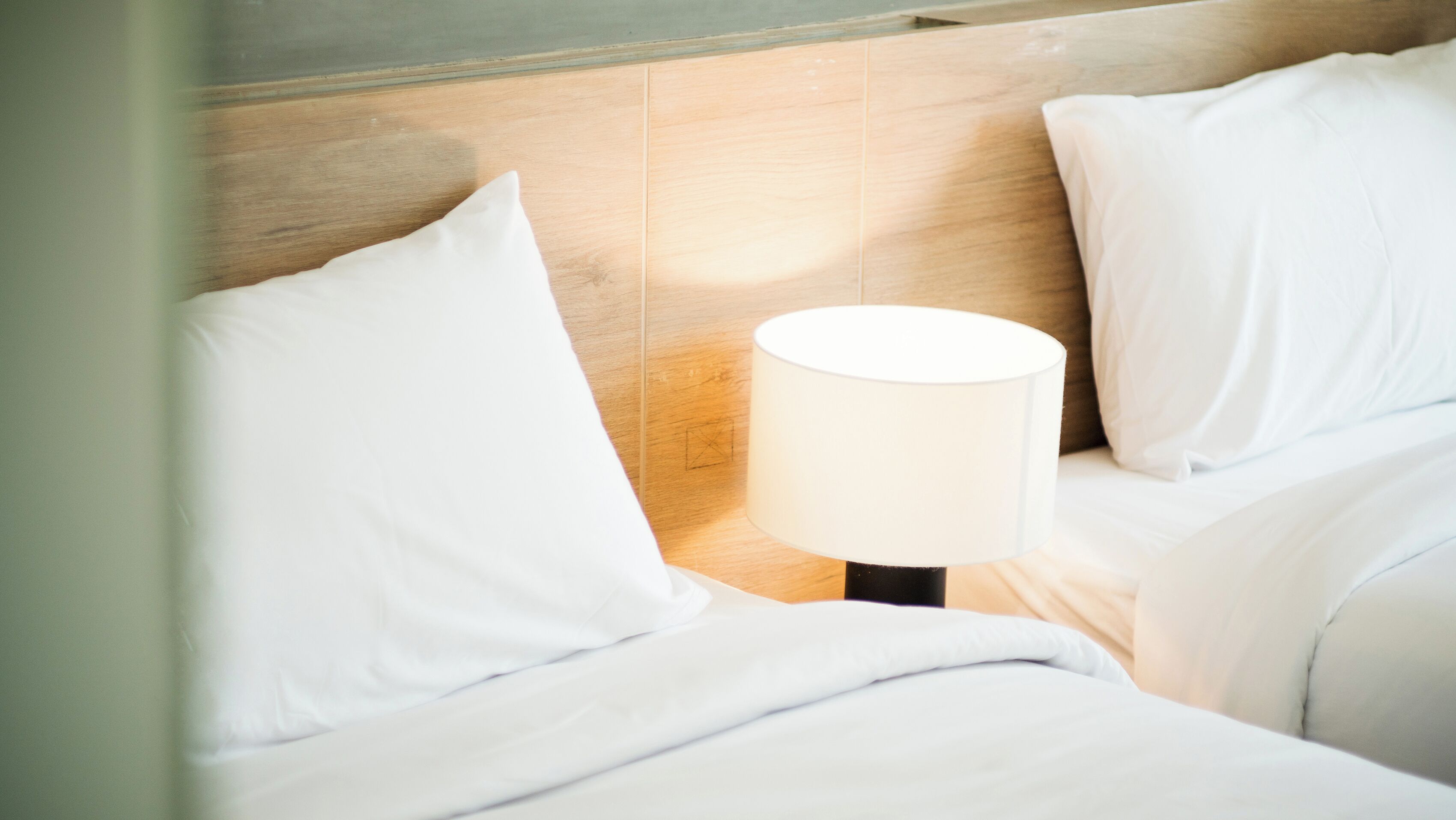 Een netjes opgemaakt bed met witte lakens en kussens, een bedlampje en een houten hoofdbord.