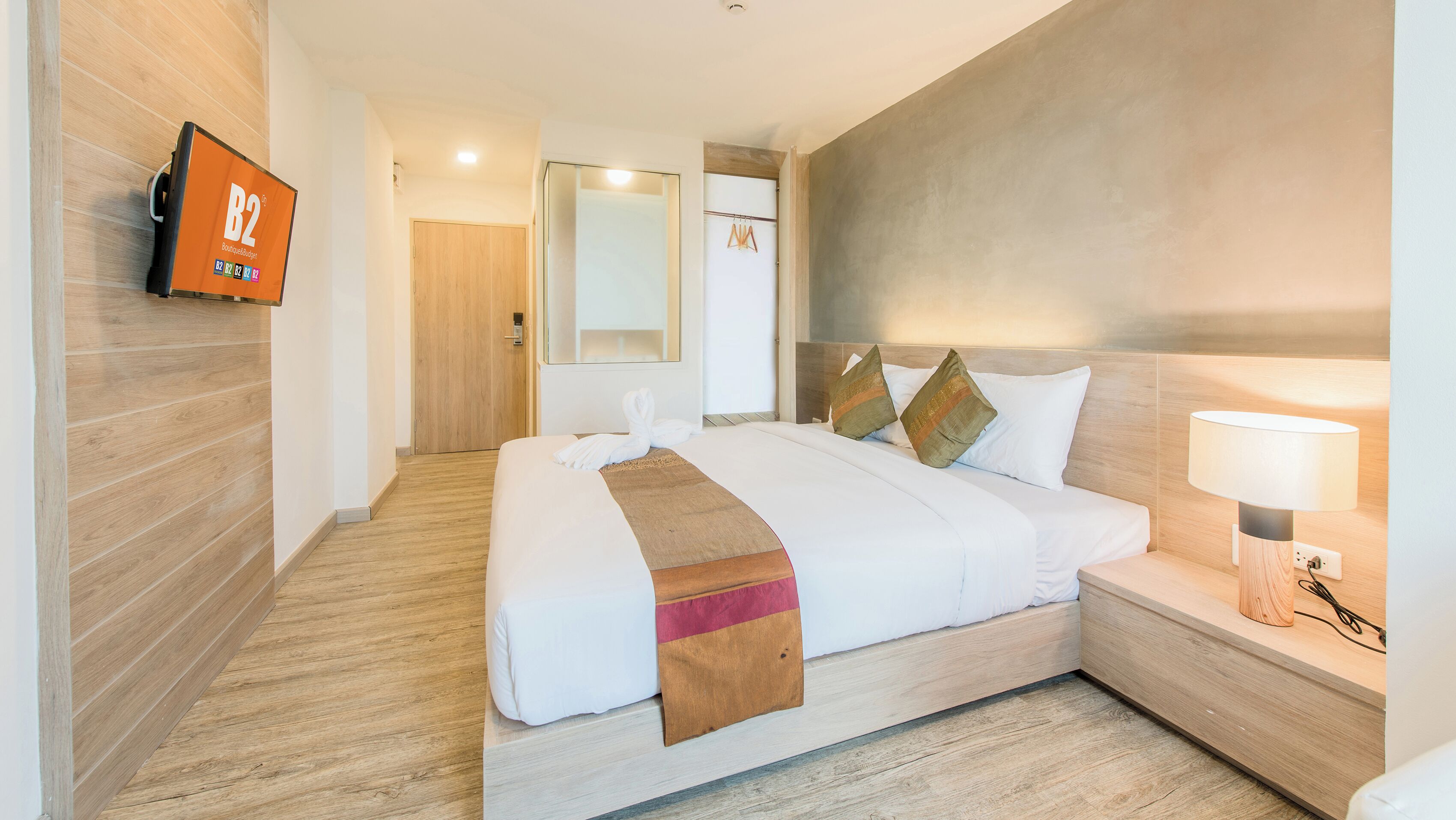 A hotel room with a bed, a bedside table with a lamp, a wooden floor, a wall-mounted TV, and a door.