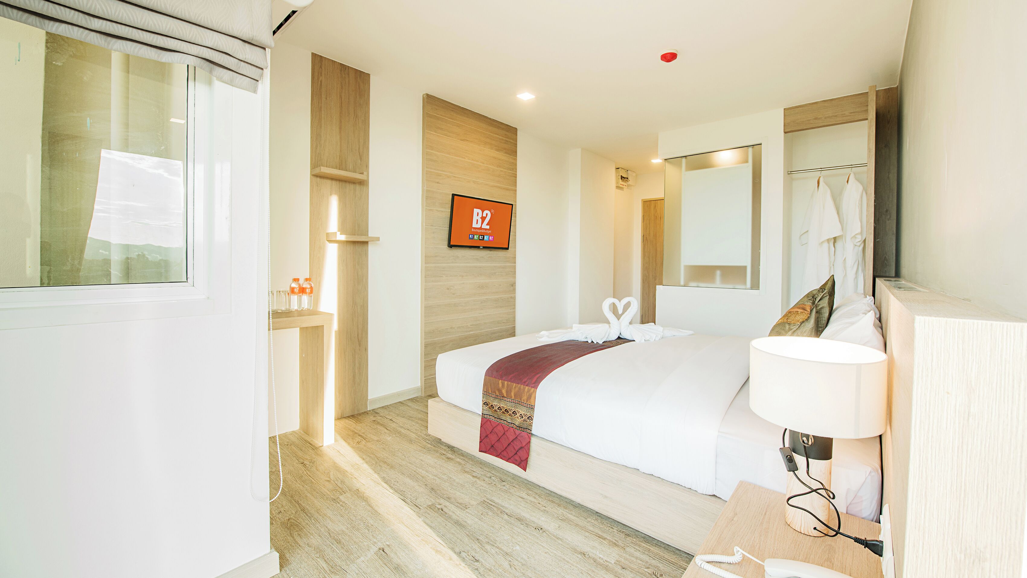 Habitación de hotel con cama, mesita de noche, lámpara y espejo. Se observa una estantería de madera con botellas y vasos, además de un cartel en la pared.
