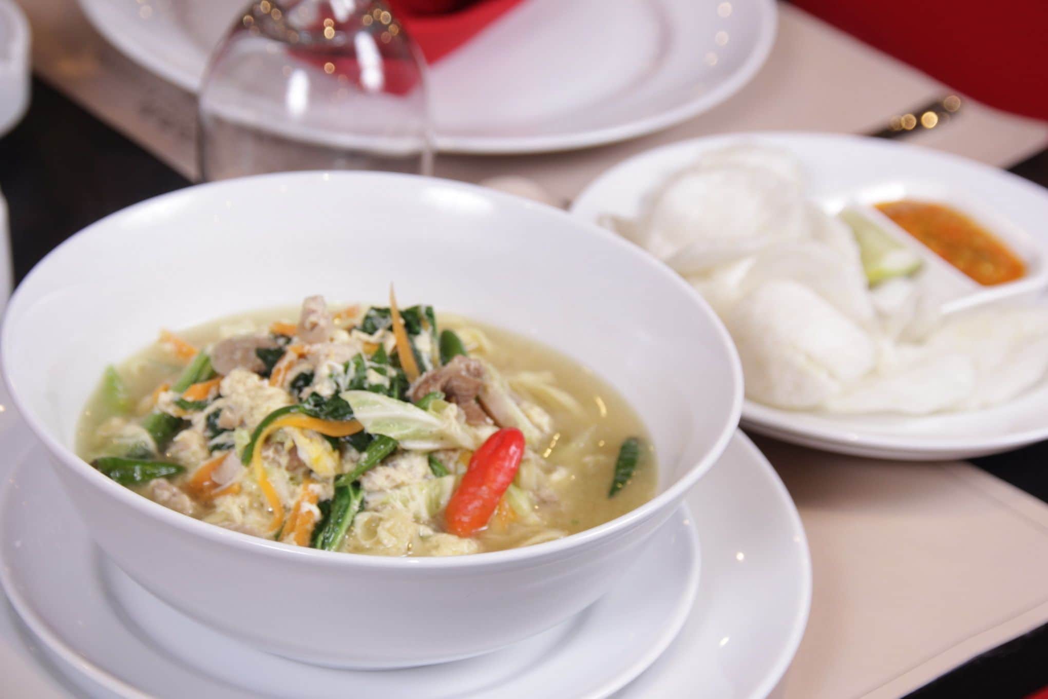 A bowl of soup with vegetables and meat, a plate of white food, and a glass of red wine on a table.