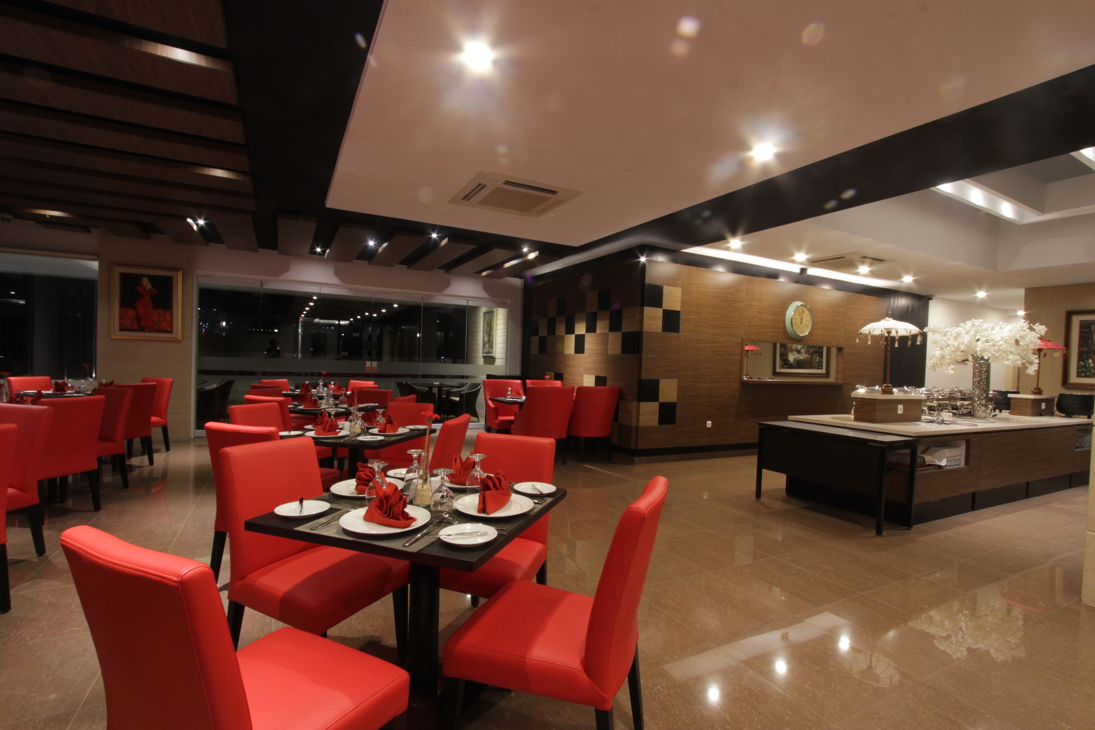 Un restaurant moderne avec des chaises et des tables rouges, un îlot central surmonté d’un lustre et une horloge au mur.