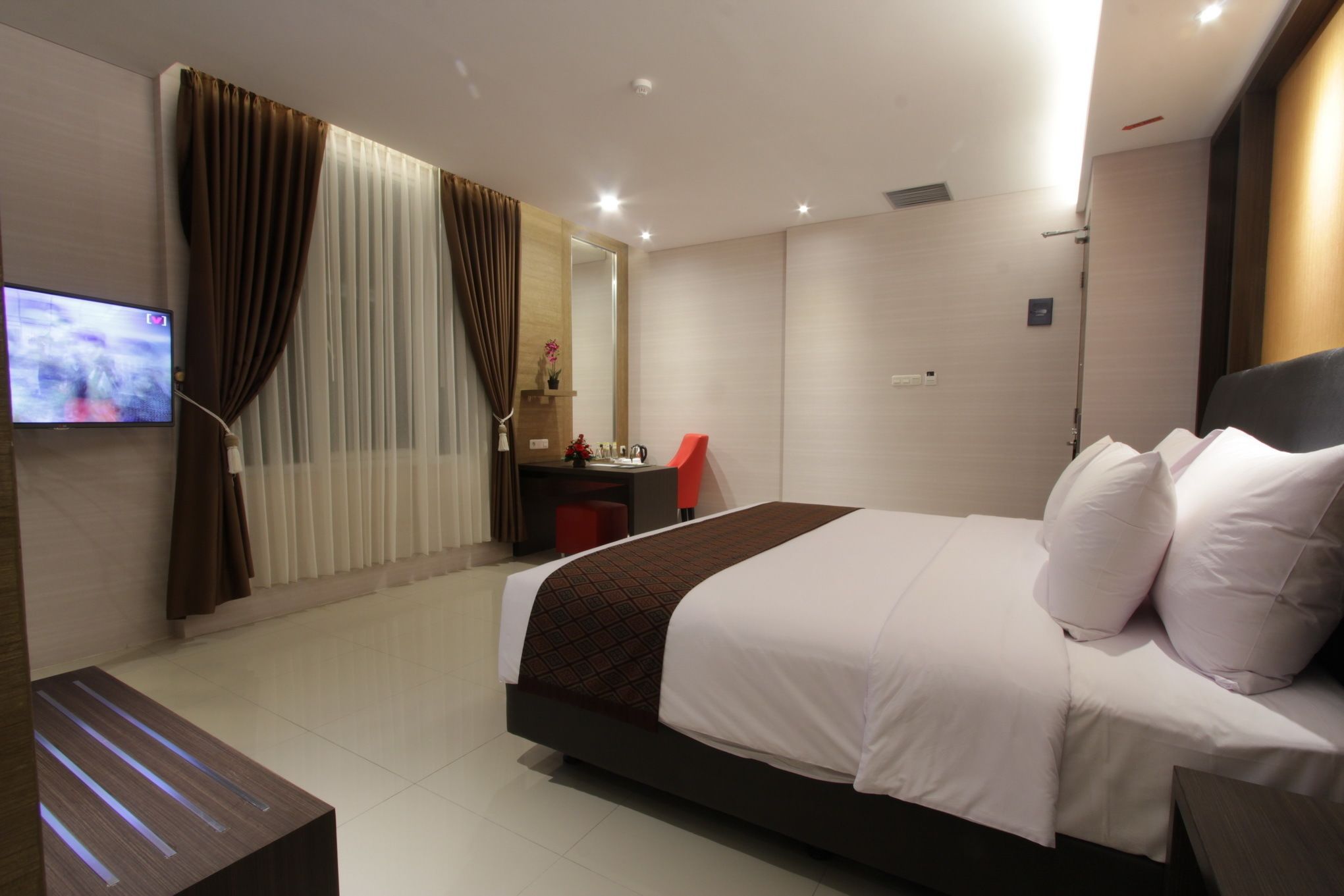 A modern hotel room with a large bed, a desk with a chair, a TV on the wall, and a window with curtains.