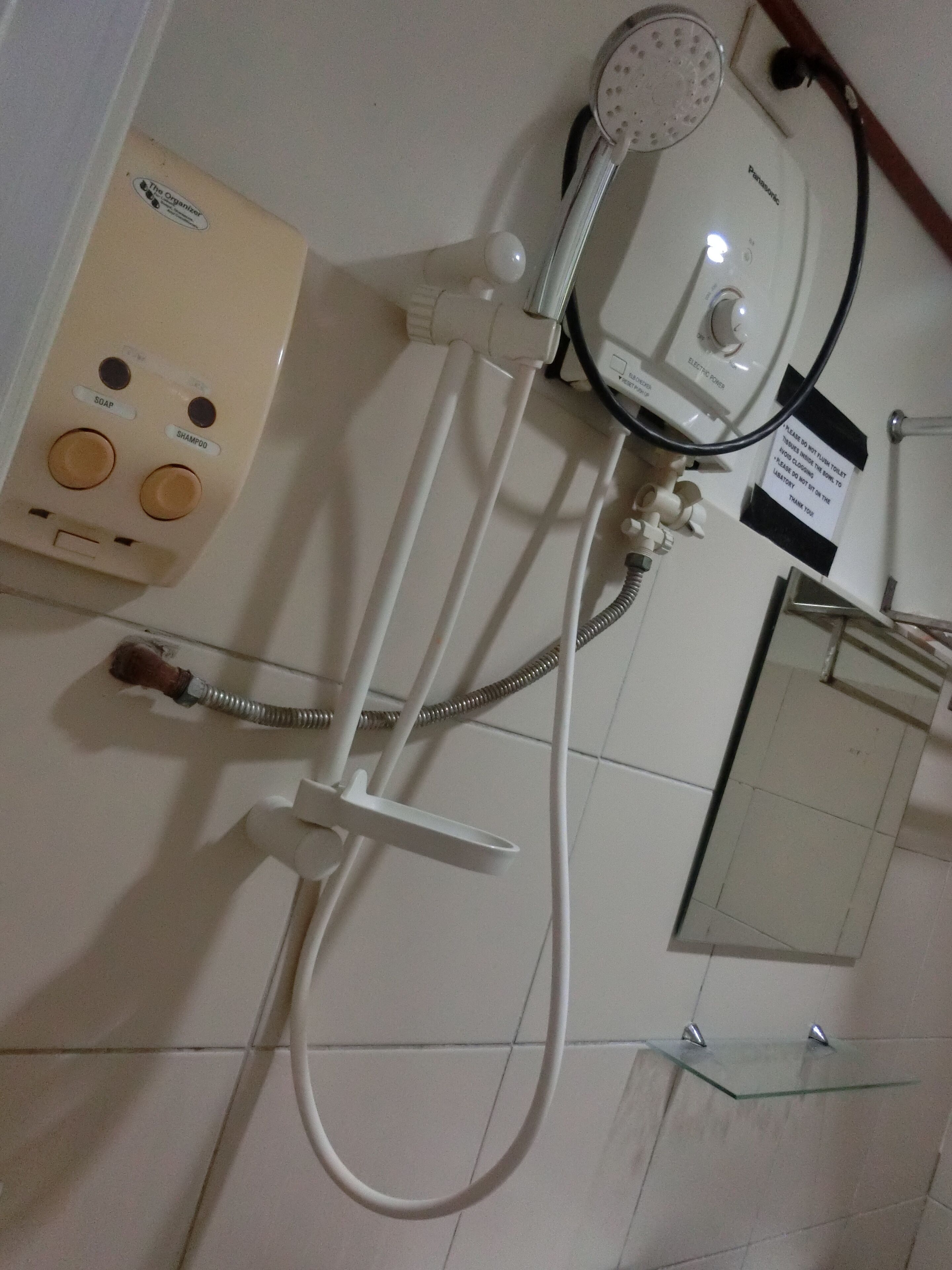 A bathroom with a wall-mounted electric shower, a soap and shampoo dispenser, and a glass shelf.